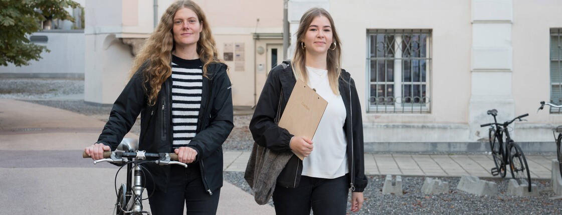 Eine Frau, die ihr Fahrrad schiebt, und eine Frau neben ihr, die ein Caritas Klemmbrett und eine Stofftasche trägt, gehen einen Gehweg entlang.