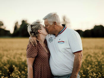 Ein älteres Paar küsst sich auf einer Wiese. Der Mann legt den Arm um die Frau.