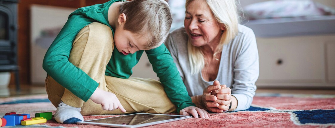 Lehrgang Fachkraft Unterstützte Kommunikation  Kind sitzt auf einem bunten Teppich in einem Dachzimmer und nutzt ein Tablet, während eine erwachsene Person daneben liegt und aufmerksam zuschaut. Neben dem Tablet liegen bunte Spielsteine, im Hintergrund ein Bett und Holzdecke.