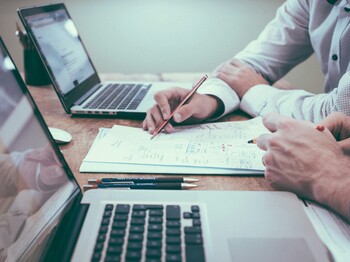 Zwei Personen arbeiten an einem Schreibtisch mit Laptops und Notizen, während sie gemeinsam Dokumente analysieren und besprechen.onto verstehen